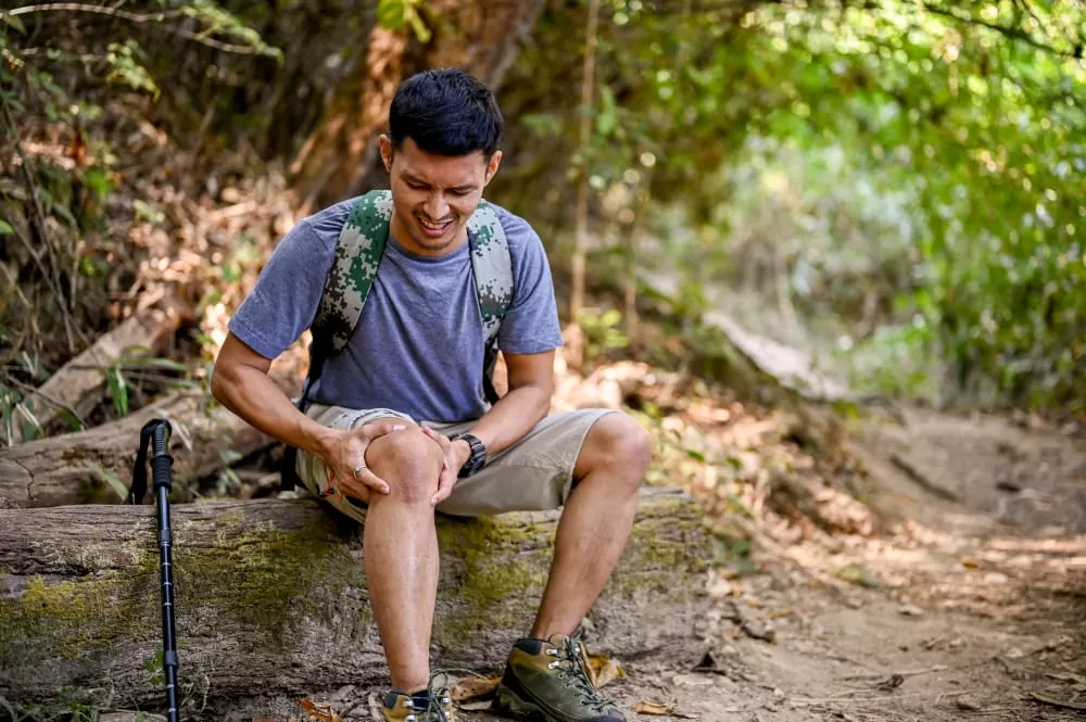 A stressed male trekker sits on a wooden log and holds his knee, suffering from knee pain